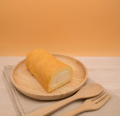Cake roll in wooden dish with wooden spoon and fork on beige wooden table,orange background,copy space