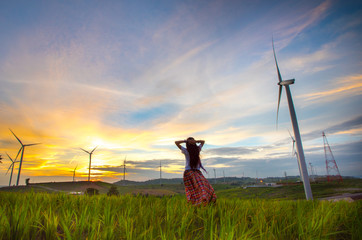 Naklejka na ściany i meble Happy woman relaxing with wind generators turbines beautiful sunset background in Khao Kho mountain, Petchabun, Thailand. Renewable energy concept.
