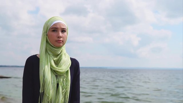 A young Muslim woman in a veil, which covers her hair, walks along the shore along the sea, the lady looks thoughtfully into the distance, she looks serious and focused