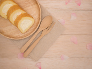 Top view cake roll in wooden plate with wooden fork,spoon  on brown fabric,beige wooden table,background with copy space,decorates with pink flower petals