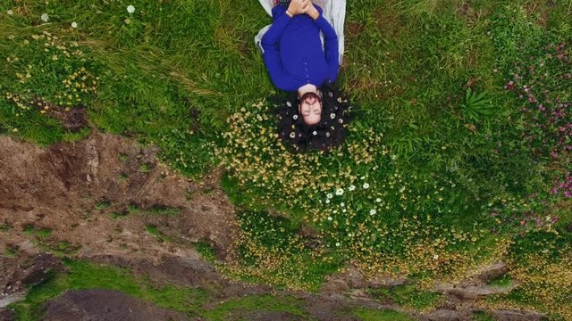 Aerial shot on peaceful tall asian adventure man smiling and laying in grass with field flowers in his long black hair looking at camera. Connection with nature power concept