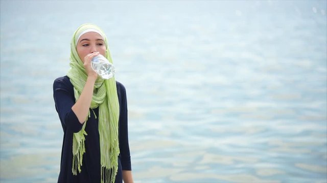 An Arab woman in beautiful hijab drinks water from a plastic bottle, a lady is near the sea or the ocean on a summer day, maybe it's hot outside