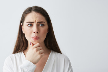 Portrait of nervous young brunette girl thinking looking in side holding hand on chin over white background.