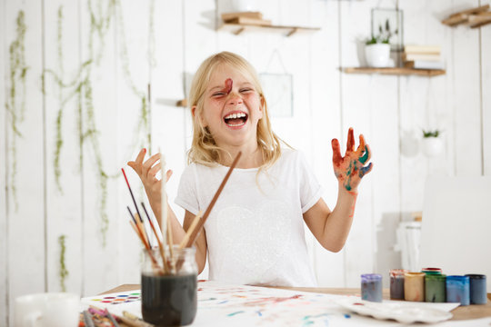 Laughing, charming little girl with blonde hair, freckles and blue eyes messed up herself with paint. Creative child with paint on her face and hands.