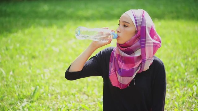 A young Muslim woman wearing a headscarf drinking mineral water from a plastic bottle in the park on a warm summer day, the lady quenches thirst