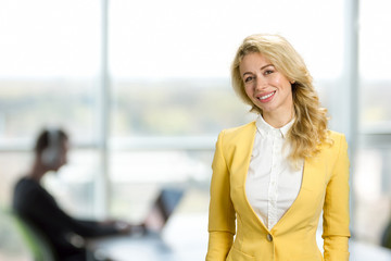 Cheerful woman on office window background. Beautiful office manager in fashion yellow jacket smiling on business center background.