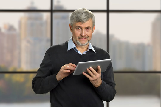 Portrait of mature man with pc tablet. Serious grey hair man holding computer tablet on office window background close up. Senior man with digital tablet on blurred background.
