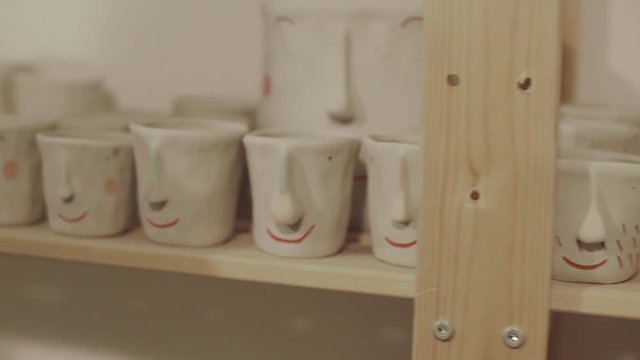 Close up view of wooden shelf full of white cute ceramic pots with faces in pottery workshop.