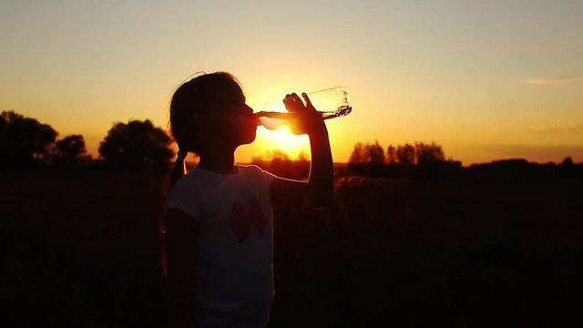 Silhouette of young girl drinking water from bottle in wheat field at sunset in summertime. Rehydrate your body.