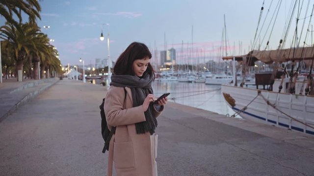 Lonely girl at early morning is texting message on her smartphone on chill peaceful background of marina harbour with many parked sail boats. She wears coat and warm scarf