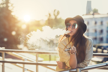 Pretty young hipster woman in black hat vape ecig, vaping device at the sunset. Toned image. - 168547896