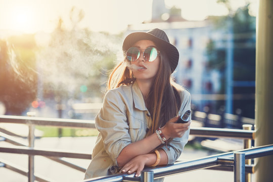 Pretty young hipster woman in black hat vape ecig, vaping device at the sunset. Toned image.