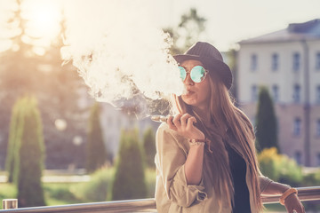 Pretty young hipster woman in black hat vape ecig, vaping device at the sunset. Toned image.