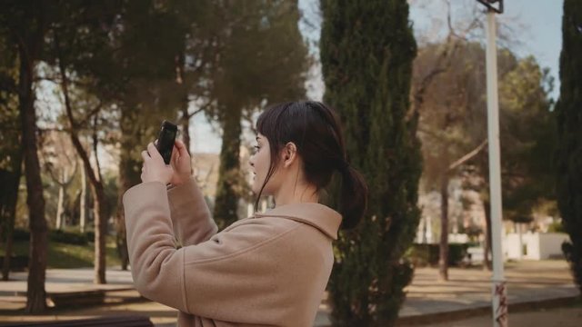 Camera moves around beautiful brunette girl , she is using application in her smartphone to take photos of something in park during her day walk