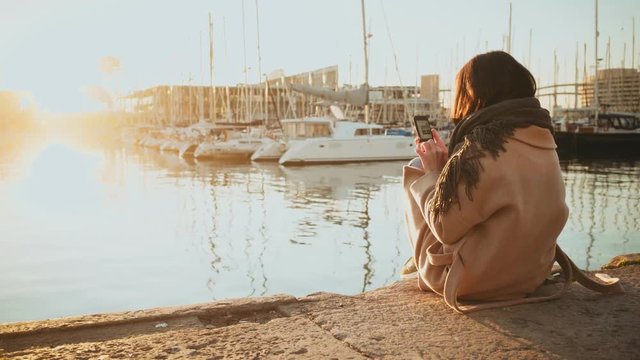 Back shot of girl in coat sitting alone on edge of pier bank in port in front of parked sail boats at early morning,texting in smartphone,then put it away and relaxing with beautiful view of sunrise