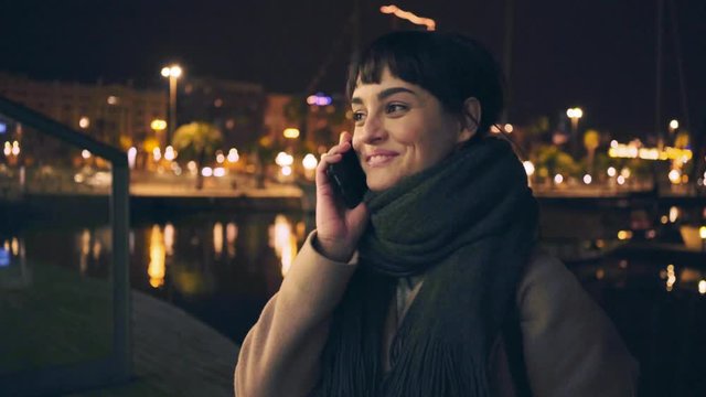 Smiling and laughing young brunette girl in coat and scarf is talking by her phone at night street in town with amber lights on backgrounds