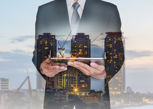 Double Exposure Of Businessman Hand Hold Tablet, Construction Crane And Building In The Evening, Twilight As Business, Technology, Communication And Industrial Concept.