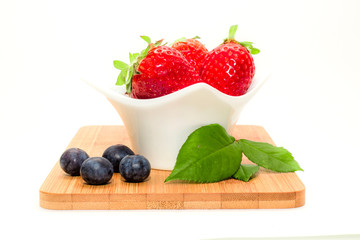Strawberries in white bowl on wooden board with blueberries and leaf
