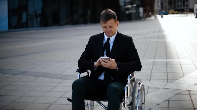 Young attractive entrepreneur business man sitting on wheelchair working with iphone outdoor. He in formal suit and tie. Confident and attractive disabled handicapped man.