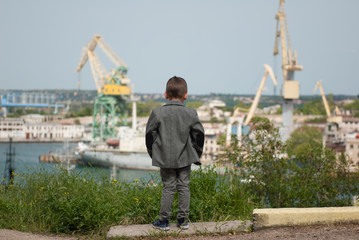 little kid in a jacket stands against the background of a sea port