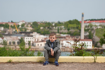 Sad little boy sitting on the curb with sea port background