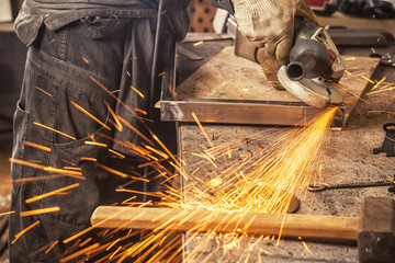 A young male welder in a black working overall and working gloves grinds a metal product with angle...