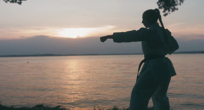 MED Caucasian professional female athlete wearing kimono practicing karate near large lake, sunset shot. 4K UHD, 60 FPS SLO MO