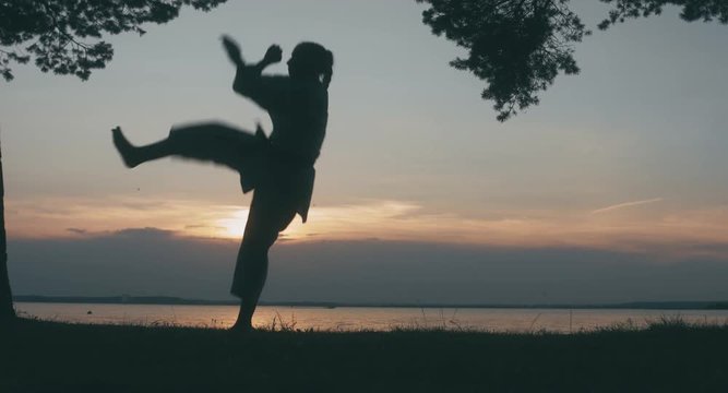 MED Caucasian professional female athlete wearing kimono practicing karate near large lake, sunset shot. 4K UHD, 60 FPS SLO MO