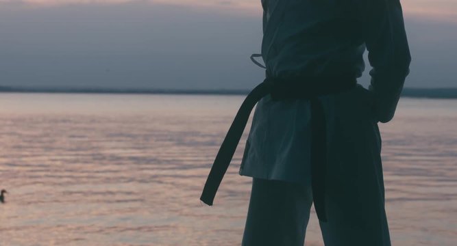 CU female karate fighter tightens the black belt on her kimono. Sunset over large lake in the background. 4K UHD, 60 FPS SLO MO