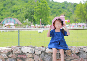 Cute little asian girl wear straw hat sitting on the stone wall against green public garden. Child girl looking out.