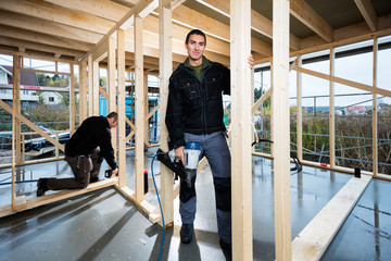 Carpenter Holding Drill While Colleague Working At Construction  - obrazy, fototapety, plakaty