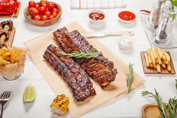 Spicy hot grilled spare ribs from a summer BBQ served with chips, corn and fresh tomatoes on an old wooden cutting board