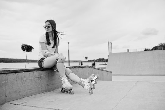 Portrait of an attractive young woman in shorts, t-shirt, sunglasses and rollerblades sitting on the concrete bench in the outdoor roller skating rink. Black and white photo.