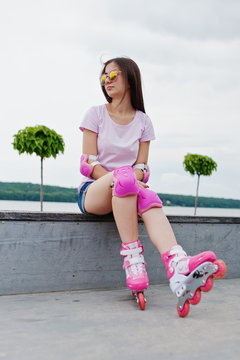 Portrait of an attractive young woman in shorts, t-shirt, sunglasses and rollerblades sitting on the concrete bench in the outdoor roller skating rink.
