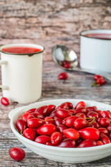 
 Ripe red dogwood fruits in a white plate, next to a mug with compote on an old wooden table. The concept of a healthy diet. 