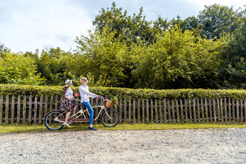 Fototapeta premium Young couple riding a bike tandem in the park. Against the background of the fence from the tree