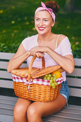 Positive redhead female sits on a bench with a picnic basket.