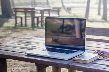 Laptop on a wood table in a public park with notebook paper and sunglasses on side.