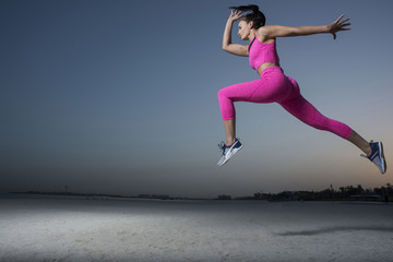 Fototapeta na wymiar A side view of a strong athletic, beautiful female sprinter as she does a running jumping with an early morning sunrise and dark silhouettes of homes wearing a tight pink fitness outfit 