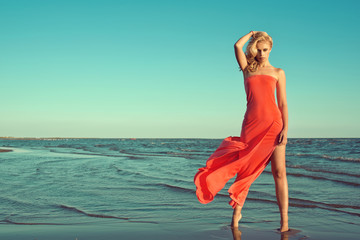Gorgeous sexy slim blond model in red strapless dress with flying train standing on tiptoe in the sea water, holding her wavy hair blown by the wind. Copy-space. Outdoor shot
