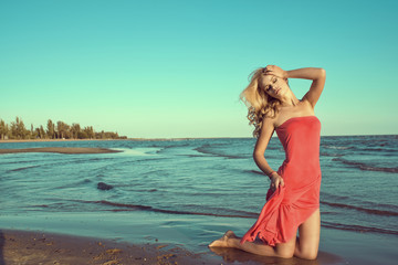 Naklejka premium Gorgeous sexy slim blond model in red strapless dress standing on knees in the sea water with closed eyes, one hand on her head, another one holding the train of her gown. Copy-space. Outdoor shot