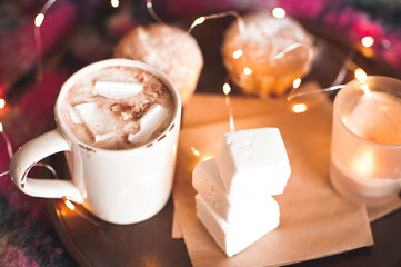 Mug with coffee and marshmallow with cake and candle stay on wooden tray over christmas lights background. Good morning. Selective focus.