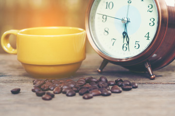 Yellow ceramic cup , roasted coffee beans  and classic alarm clock on wood table