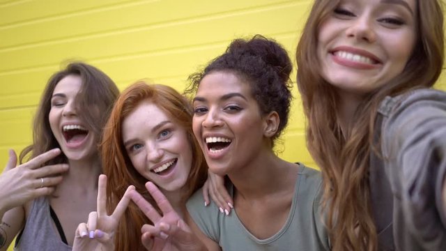 Four multiethnic beautiful female friends making selfie and showing different emotions near yellow wall