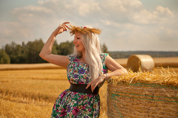 beautiful rural woman wearing flower dress and wheat crown standing near dry round straw haystack...