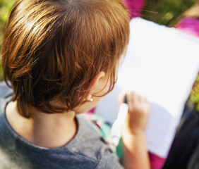 Pretty beautiful child girl sitting on the grass in the park and make notes in the notebook. The girl working, studying, learning, keeping a diary.