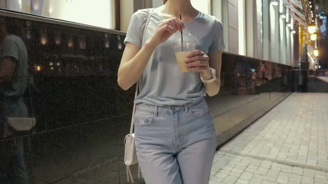 Young woman in jeans and a T-shirt is drinking a cocktail at night in a summer city