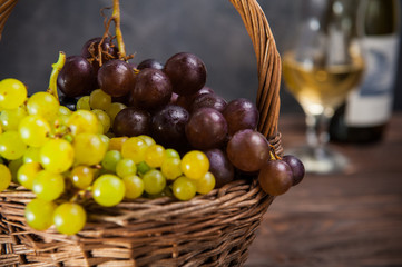 Wicker basket with various grapes: red, white and black berries on the dark wooden table with bottle and glass of white wine in the background.. Selective focus. Copy space