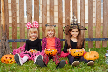 Youthful girls in halloween costumes sitting on grass by fence and eating treats