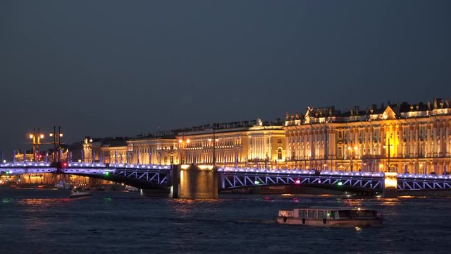 Pleasure boats sail under the palace bridge in St. Petersburg at night. In the background, the Hermitage